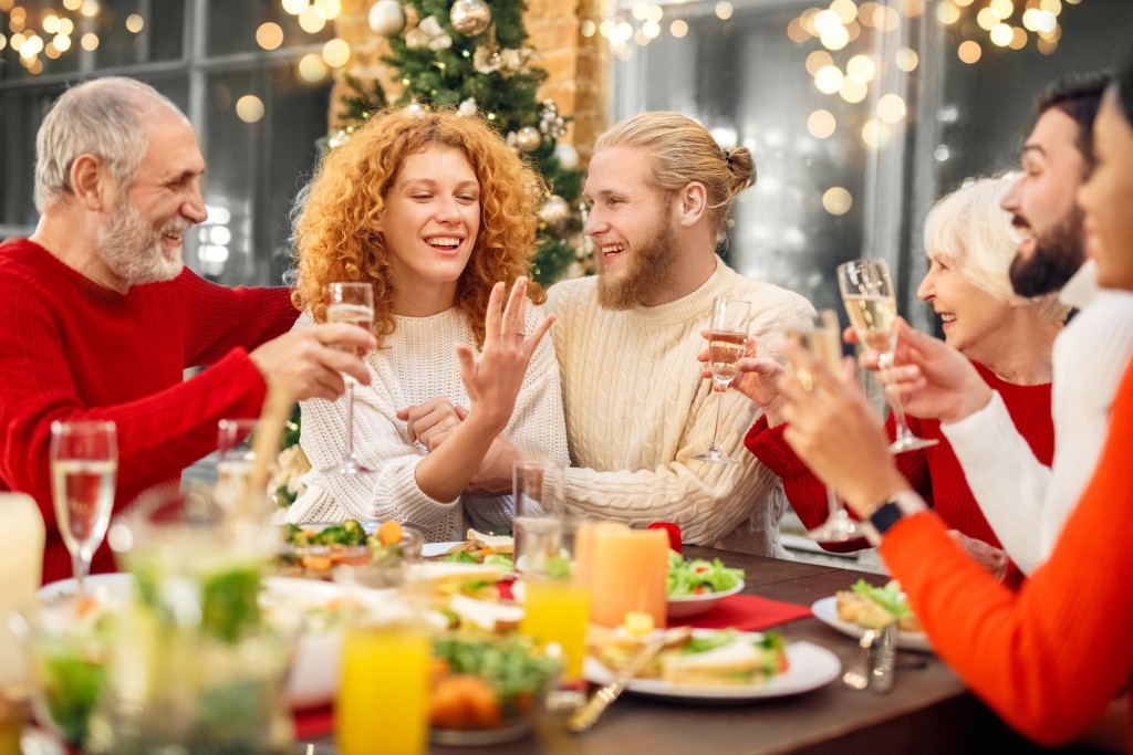 Happy woman sitting behind table, showing wedding ring on finger to the relatives. Couple celebrating engagement together with family